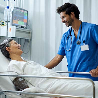 A male nurse is standing at the bedside of his hospitalized patient, smiling, and looking attentively. The patient is reclining and is attached to a blood pressure monitor and a nasal cannula.