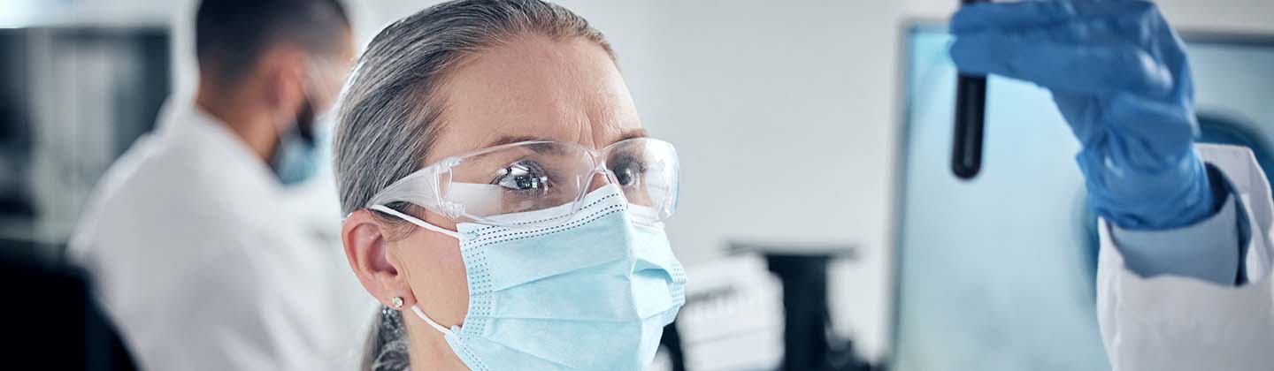 A female nurse is wearing a lab coat, protective eyewear, mask and gloves, and she is holding up and inspecting a blood specimen in a tube.