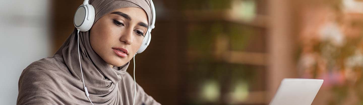 A focused nursing student in a hijab is studying for the NCLEX exam at a desk. She is wearing headphones and looking intently at her laptop screen while taking notes. A coffee cup and various study materials are organized around her, suggesting a productive study session in a quiet, indoor environment.