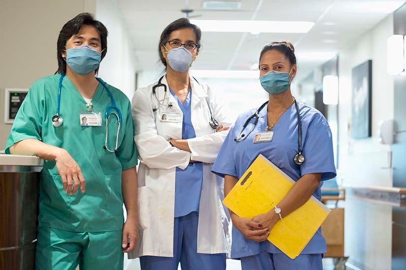 A diverse team of nurses gathers around a table for a meeting, with one standing and leading the discussion, illustrating collaboration and engagement in a clinical setting. 