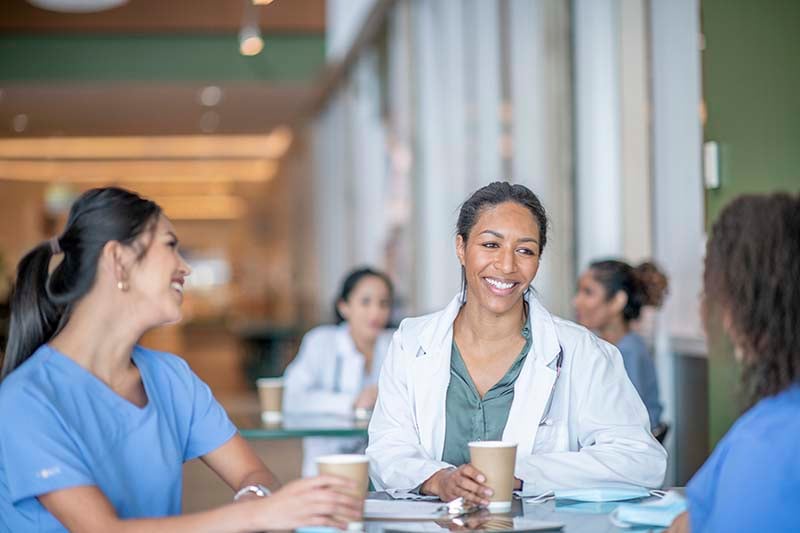 A group of nurses are in a medical center cafeteria seated at a table. They are smiling and laughing and engaged in a pleasant conversation.