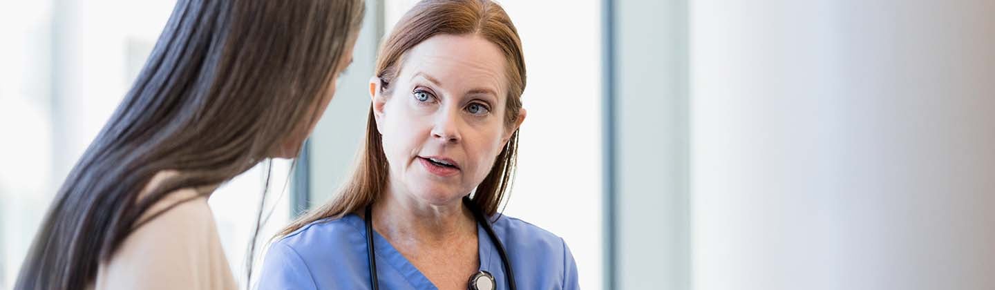 A nurse in blue scrubs with a stethoscope around her neck is attentively listening to a patient while holding a clipboard. They are standing in a brightly lit room with a large window in the background.