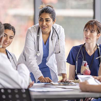 A group of attentive nurses in scrubs and white coats gather around a table for a team briefing, led by a nurse standing up, indicating a collaborative medical discussion.