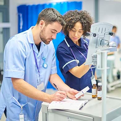 Two nurses are standing at a medication cart in the Recovery Room. A male nurse is holding a box of medication and doing documentation in a logbook. A female nurse is verifying the correct medication and entering information on a computer screen.