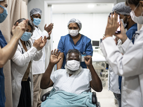 An elderly male patient is being wheeled out of the hospital and hospital staff have lined the hallway and are clapping in happiness for his recovery.