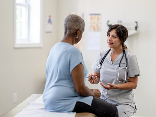 A patient is seated on an exam table in a medical office. A nurse is seated on a chair next to the patient, and they are both engaged in conversation with each other.