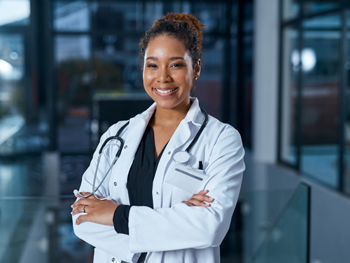 A smiling female nurse wearing dark scrubs and a white lab jacket has her arms crossed in front of her and is confidently facing forward.