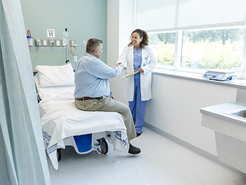 A female nurse practitioner is meeting with a clinically obese male patient being discharged from the hospital. They are shaking hands and smiling as the nurse practitioner is discussing his discharge plan.