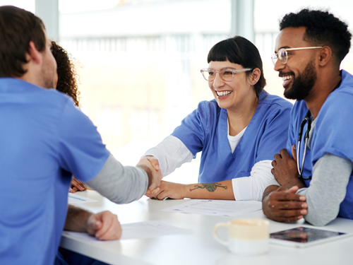 A group of nurses wearing blue scrubs are seated around a conference table and smiling. Two nurses are shaking hands across the table.