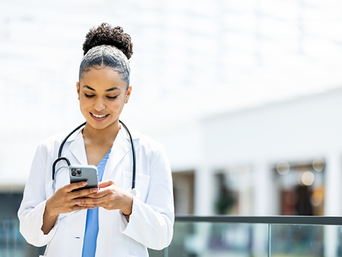 A young female nurse wearing light blue scrubs and a white lab jacket is standing in a building atrium and smiling while looking at information on her cell phone.